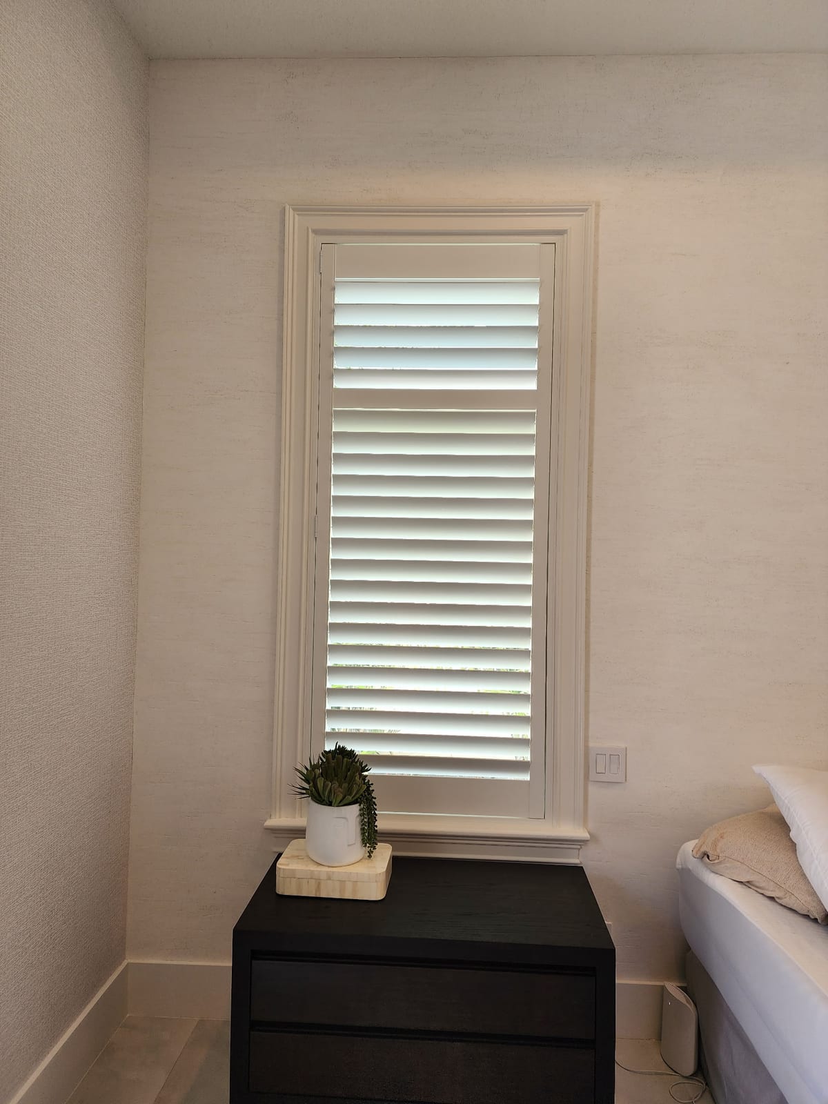 A white shutter on a bedroom window above a bedside table