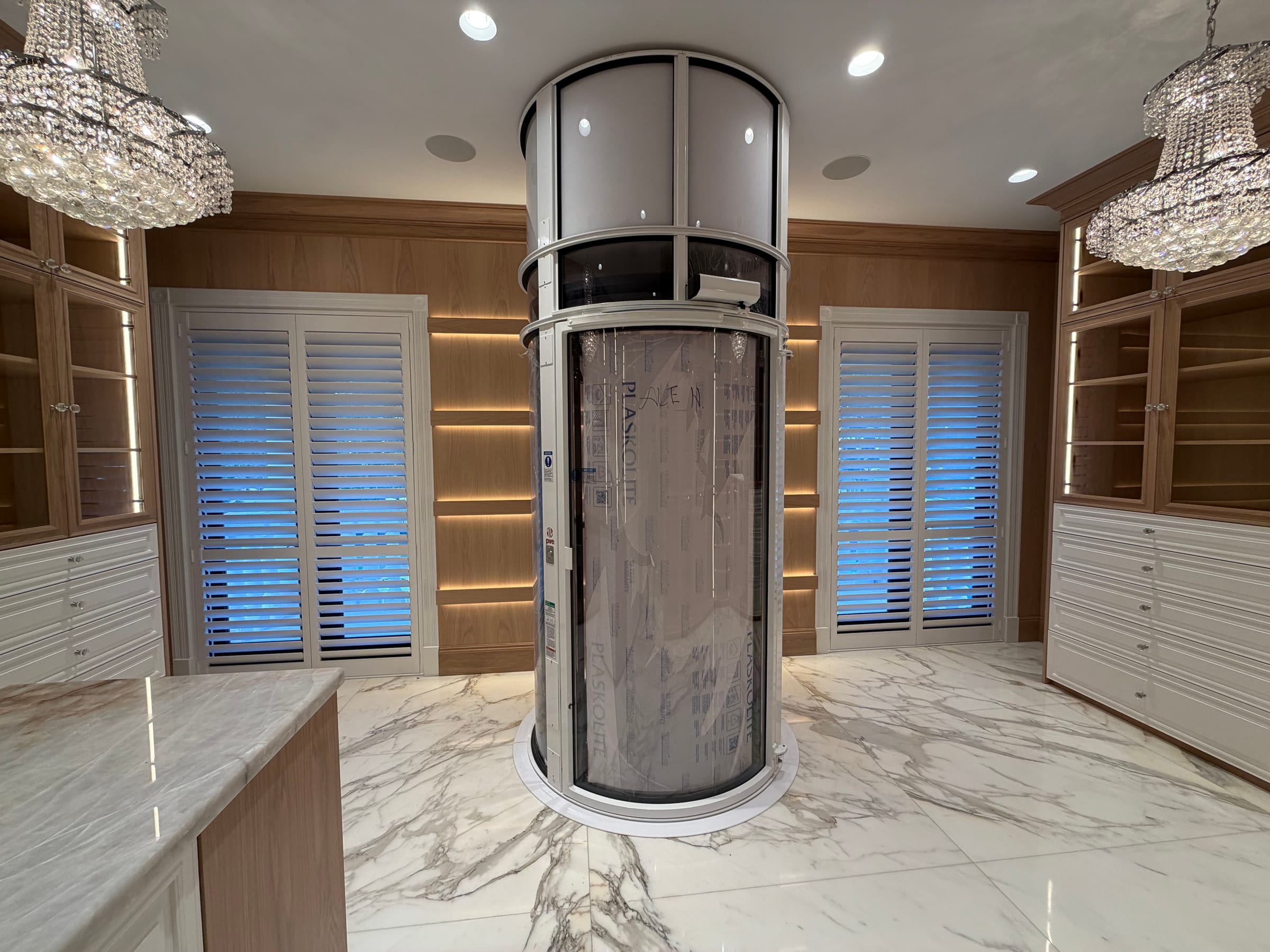 White shutters on tall windows in a panelled room with crystal chandeliers, beside a glass lift