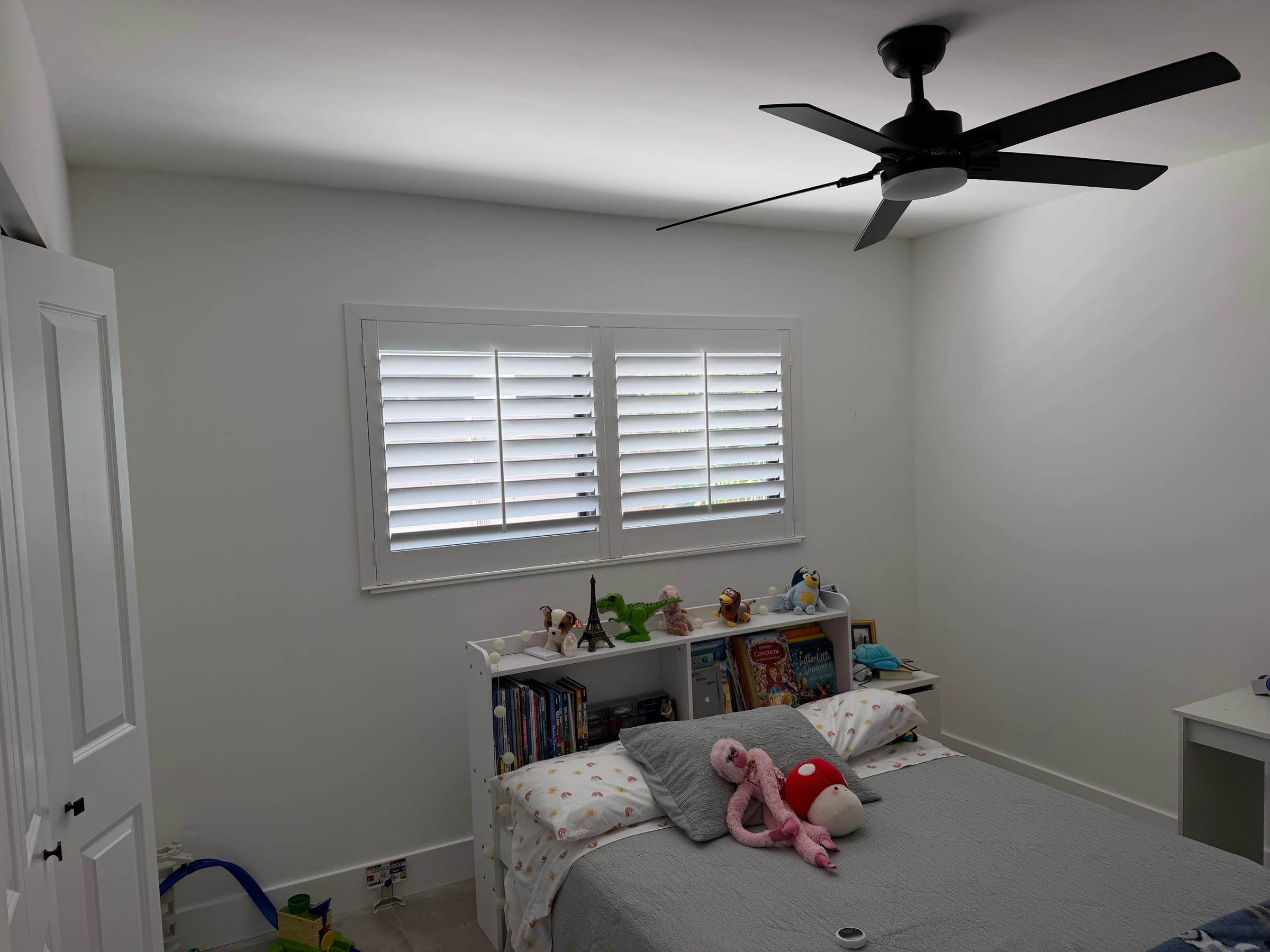 White shutters on a bedroom window above a low bookcase and a made bed