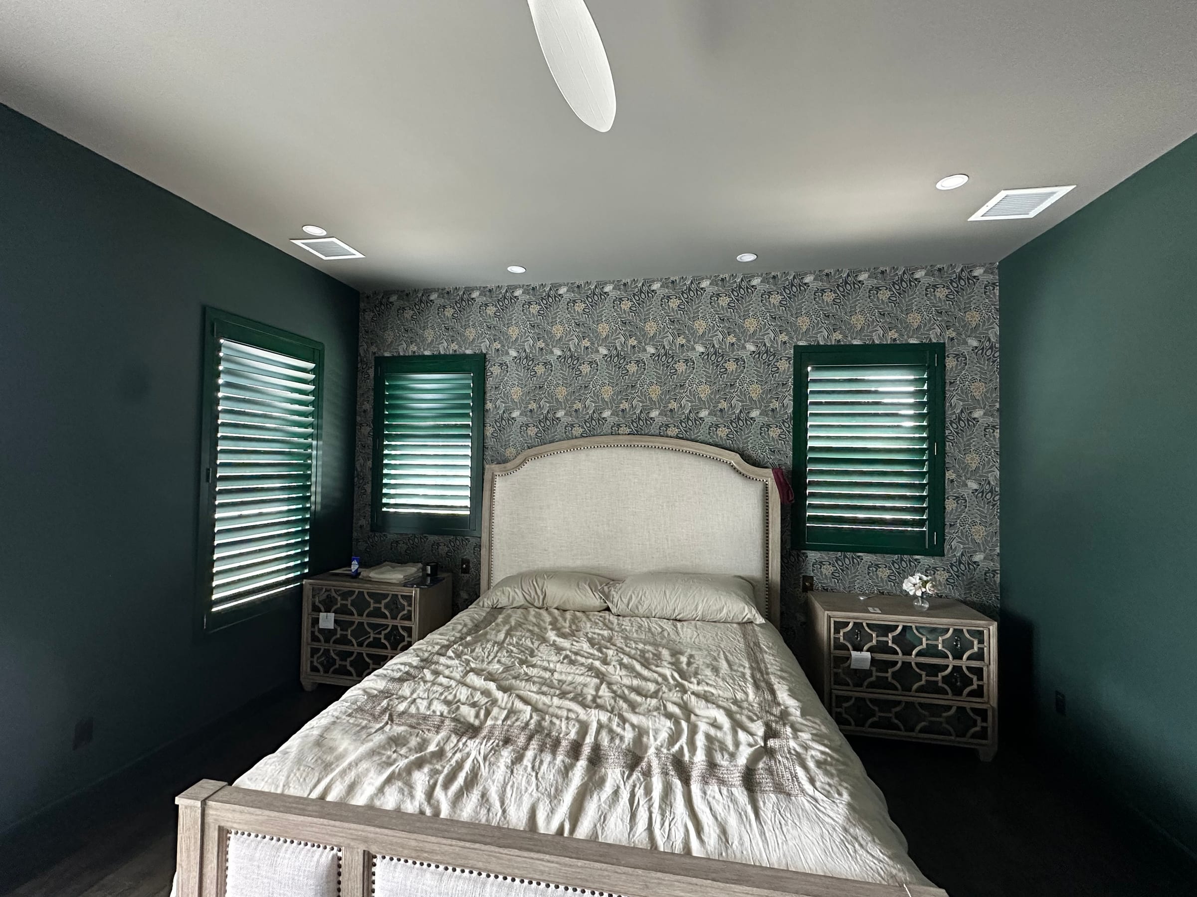 Green-painted shutters on bedroom windows either side of a floral papered wall